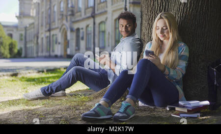 Mann mit Handy sitzen unter Bäumen und Blick auf Mädchen mit Telefon, Zuneigung, Filmmaterial Stockfoto