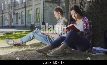 Mann mit Laptop unter Baum in der Nähe von Mädchen lesen Buch sitzen, zeitgenössische Jugend Stockfoto