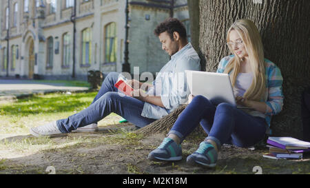 Junge männliche sitzen unter Baum mit Buch in der Nähe von Frau mit Laptop, Studentisches Leben Stockfoto