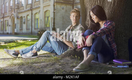 Zwei junge Menschen sitzen unter Baum, Mädchen unten und schauen Kerl bei Ihr zu suchen Stockfoto