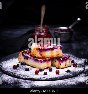 Stücke von Quark Torte mit blaubeere Beeren mit Zucker bestreut Pulver, hinter dem Glas mit Marmelade, schwarzer Hintergrund Stockfoto