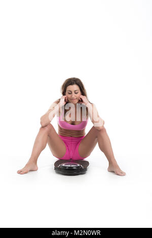 Junge Frau sitzend vor Skala unglücklich mit Ihrem Gewicht Gestik Traurigkeit. Fit und gesund. Auf weissem Hintergrund Stockfoto
