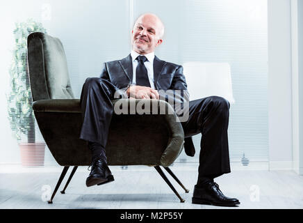 Entspannt erfolgreicher Geschäftsmann mit einem zufriedenen Lächeln in einem Sessel sitzend mit einem Bein baumelnd über die Seite in die Kamera grinsend Stockfoto