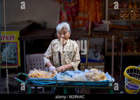 Ältere asiatische Frau. Thailand Straßenverkäufer. 84-jährige Frau, die Lebensmittel aus einem Wagen verkauft. Thailändischer Straßenmarkt. Stockfoto