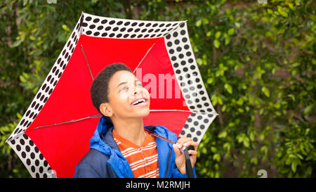 Niedliche happy African American oder schwarzen Jungen mit roten Dach und trägt blaue Regenjacke. Lachen, Lächeln, Junge schaut hoch am Himmel. Kopieren Raum auf der rechten Seite Stockfoto
