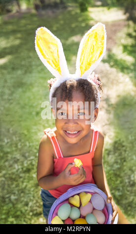 Nette und schöne afrikanische amerikanische Mädchen mit Hasenohren, isst mit Freude Ostern marshmallow Candy während Ostern Korb voller Eier. Mädchen sitzen Stockfoto