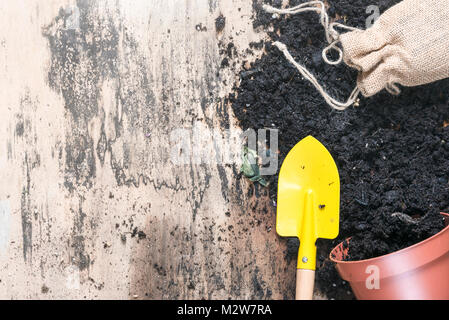 Blumenerde Pflanzen im Frühjahr Konzept mit einem Blumentopf umgeworfen, voll mit Erde, ein Garten Spaten und einem Grundgewebe aus Jute Sack, auf einem vintage Holz- Arbeitsbereich. Stockfoto