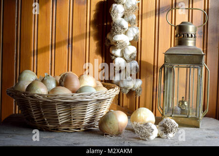 Zwiebeln auf einen Korb, Knoblauch Zopf hängen auf Holz Wand und eine Öllampe Stockfoto