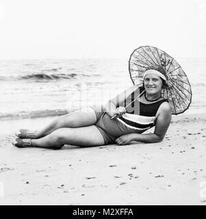 Junge Frau erstreckt sich am Strand mit ihrem Schirm, Ca. 1924. Stockfoto