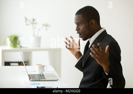 Schockiert afrikanisch-amerikanischen Geschäftsmann im Anzug Gefühl durch on-line-Nachrichten am Bildschirm des Computers am Arbeitsplatz mit Laptop suchen betäubt, betonte Trad Stockfoto