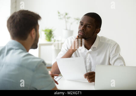 Ernsthafte aufmerksam afrikanischen hr manager Hören kaukasischen Kandidat im Vorstellungsgespräch, konzentrierte sich strenge Arbeitgeber denken in Zweifel über die Einstellung Dez Stockfoto