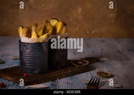 Horizontale Foto von zwei Portionen gebratenen Kartoffeln Streifen mit der Haut. Hausgemachte Pommes Frites sind in alten Vintage Dosen mit Papier im Inneren und auf Platziert abgenutzt Stockfoto