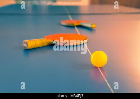 Sportliche Wettbewerbe im Tischtennis. Zwei alte orange Tennisschläger und eine gelbe Kugel liegen auf einem blauen Tischtennisplatte Stockfoto