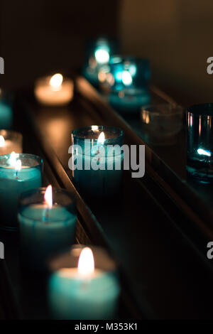 Lit votive Kerze in türkis blau Inhaber auf dunklem Metall Regal bei schwachem Licht. Stockfoto