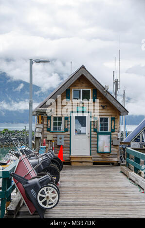 Haines, Alaska, USA - 6. Juli 2017; Holzsteg mit Schubkarren zu Hafenamt led gesäumt am Wasser mit Wasser, Berge und bewölkt. Stockfoto