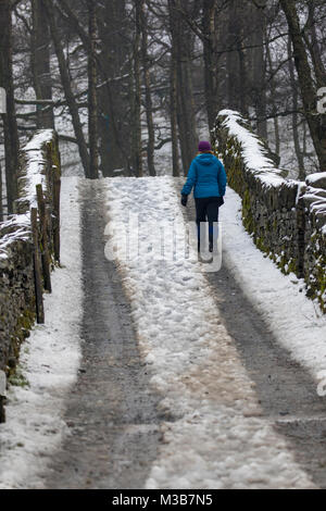 Eine einsame weibliche Walker laufen über eine Brücke in Erschliessung Dorf Chapel Stile der Great Langdale Beck Kreuzung im Winter mit dicken Schnee auf dem Boden, Great Langdale, Cumbria, Großbritannien Stockfoto