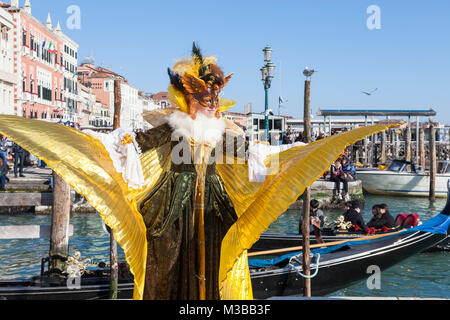 Venedig, Venetien, Italien, 10. Februar 2018. Bunte Kostüme beim Karneval in Venedig an einem sonnigen Tag am letzten Wochenende des Festivals. Frau in ein klassisches Kostüm mit ausgebreiteten Gold Flügel am Wasser in San Marco mit einer Gondel mit Touristen hinter ihr. Stockfoto