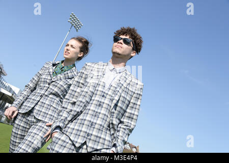 Ein schönes Mädchen mit einem Maßanzug und einen stattlichen gemischten Rennen Kerl in einem Anzug Spaziergang um einen Cricket Ground an einem hellen Tag Stockfoto