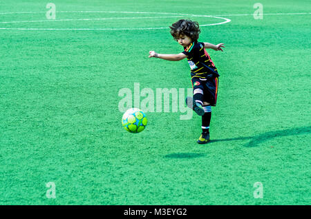 Ein niedliches, Curly schwarzhaarige Junge tritt ein Fußball auf ein Feld der grüne Rasen. Stockfoto