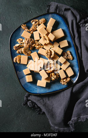 Gesalzene caramel fudge Bonbons auf blau Keramik Platte mit karamellisierten Walnüssen auf Textil Serviette über schwarzen Textur Hintergrund serviert. Ansicht von oben, Platz. Des Stockfoto