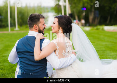 Lächelnd Hochzeit paar von der Rückseite im Freien. Bräutigam und Braut umarmen und Lachen in den grünen Park sitzen auf dem Gras Stockfoto