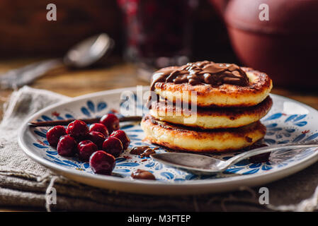 Stapel von Quark Pfannkuchen mit Haselnusspaste, gefroren Cherry und Vanilla Pod. Teekanne mit Löffeln und Glas von Beeren für den Hintergrund. Stockfoto