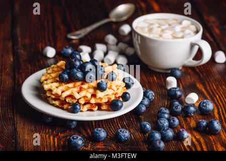 Belgische Waffeln auf dem Teller mit frischen Heidelbeeren und Tasse heißen Kakao mit Marshmallows. Löffel und einige Beeren und Marshmallows verstreut auf hölzernen Tisch Stockfoto