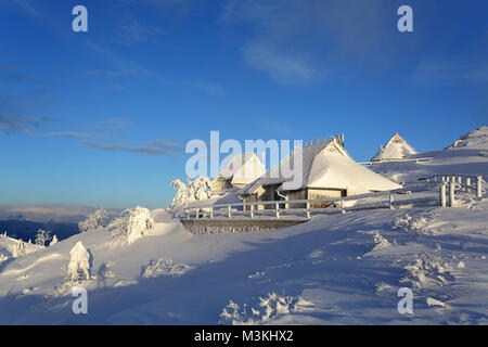 Idyllische gefroren alten hölzernen Almhütten mit Schnee in der Berglandschaft von Velika planina, Slowenien bei Sonnenuntergang abgedeckt. Stockfoto