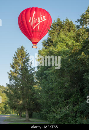 Eine Jungfrau Heißluftballon fliegen tief über ein Warwickshire Village auf der Suche nach einem Ort zu landen. UK. Stockfoto