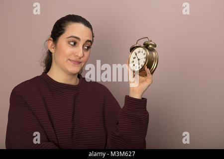 Junge Frau zweifelhaft an einem Wecker mit einem Blick der Kontemplation suchen, wie sie es hält in ihrer Hand Stockfoto