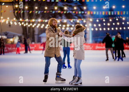 Thema Eisbahn und Verliebten. treffen junge, stilvolle Leute fahrt von Hand in Masse auf Stadt Eisbahn von Lampen und Leuchten. Eis Stockfoto