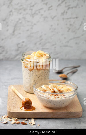 Gesunde Ernährung Frühstück. Übernachtung Hafermehl mit Chia Samen, Bananen, Peanut Butter, Honig in einem Glas auf einem grauen konkreten Hintergrund Stockfoto