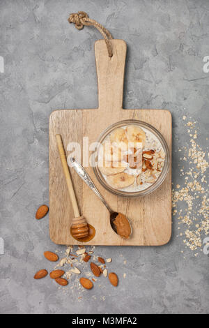 Gesunde Ernährung Frühstück. Übernachtung Hafermehl mit Chia Samen, Bananen, Peanut Butter, Honig in einem Glas auf einem grauen konkreten Hintergrund. Ansicht von oben Stockfoto