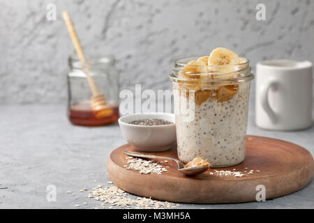 Gesunde Ernährung Frühstück. Übernachtung Hafermehl mit Chia Samen, Bananen, Peanut Butter, Honig in einem Glas auf einem grauen konkreten Hintergrund Stockfoto