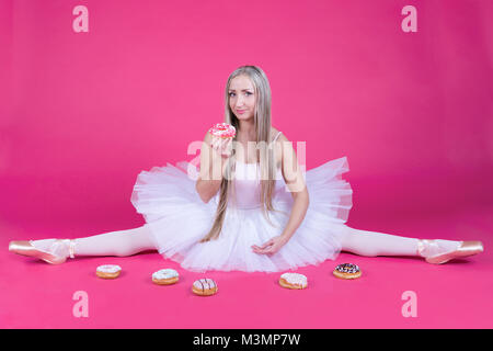 Hübsche Blondine Ballerina in Tutu Rock ein Split und essen einen Donut auf rosa Hintergrund. Stockfoto