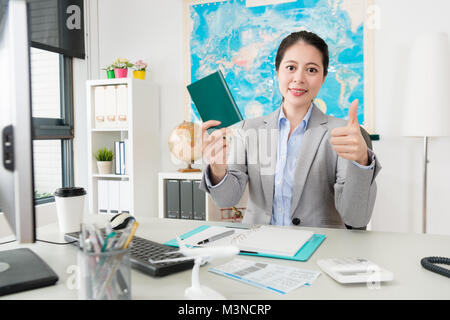 Lächelnd hübsche Frau Manager angezeigt "Daumen hoch" Geste mit Reisepass in die Kamera schauen, als Sie im Reisebüro Büro zu sitzen. Stockfoto