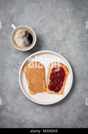 Zwei Toast mit Erdnussbutter und Marmelade, eine Tasse Kaffee auf grauem Beton Hintergrund. Ansicht von oben Stockfoto