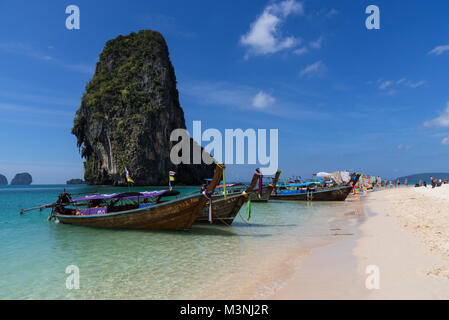Atemberaubende Ausblicke auf die Landschaft aus einer Reihe von Long-tail Boote auf einem schönen goldenen Sandstrand in Thailand mit blauem Himmel verankert und hoch aufragenden Felsen Insel Stockfoto