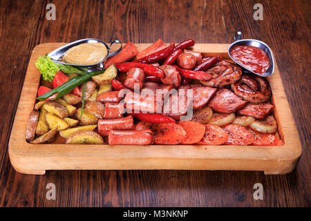 Auf dem Tisch ist ein Holzbrett. an Bord - Verschiedene gebratene Würstchen, gegrillte Karotten, Zwiebeln HaGelilit, gegrillte Kartoffeln, Soße Adjika, Senf Sauce Stockfoto