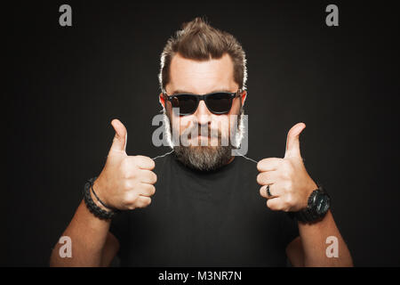 Ein schöner, starker Mann mit einem stilvollen Frisur und Bart zeigt zwei Daumen hoch im Studio auf einem schwarzen Hintergrund. Kopieren mit Platz für Ihren Text. Stockfoto