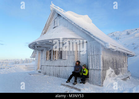 Mutter und Sohn sitzen vor der Haustür eines gefrorenen alte Holzhütte in eine verschneite Berglandschaft, Velika Planina, Slowenien. Stockfoto