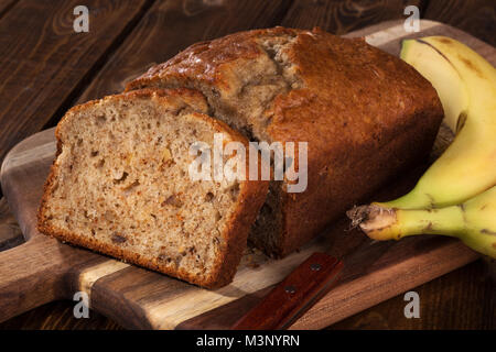 Nahaufnahme von einer Scheibe Banane Mutter Brot auf einem hölzernen Schneidebrett mit zwei Bananen auf der Seite Stockfoto