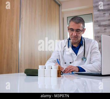 Doktor der Medizin saß an seinem Schreibtisch und eine Vorschrift schreiben oder etwas Papier arbeiten. Der Arzt im weißen Kittel mit einem Stethoskop um den Hals. Stockfoto