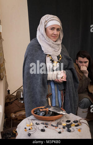 York, UK, 12. Februar 2018. Frau gekleidet wie ein Wikinger auf der jährlichen Jorvik Viking Festival. Sie ist die Teilnahme an einem realistischen Re-enactment von Leben bei einem Viking Markt & steht ein open-air abgewürgt. Auf dem Tisch liegt, ist eine Anzeige der Waren, die Sie verkaufen - Schmuck aus hauptsächlich von schwarzen Jet, sondern auch Bernstein und Fossilien. Ein Mann, der ist auch im Kostüm hinter ihr sitzen. Stadtzentrum von York, North Yorkshire, England, UK. Credit: Ian Lamond/Alamy Live Newscraft Stockfoto