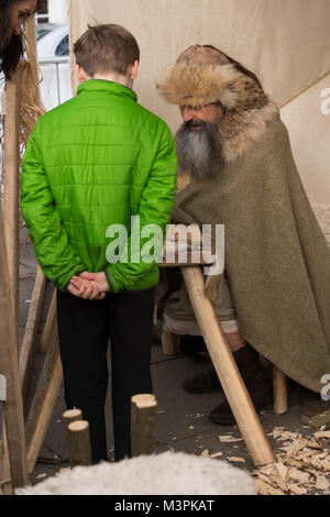 York, UK, 12. Februar 2018. Lange - der Mensch als Viking bei der jährlichen Jorvik Viking Festival gekleideten Bärtigen. Das Sitzen auf einem Stuhl, er spricht mit einem Jungen (Rückansicht) und seiner Mutter (Kopf nur sichtbar) erklärt das Runenalphabet, auf Runen auf einem Holzbrett geätzt. Er beteiligt sich an einem realistischen Re-enactment von Leben bei einem Viking Markt. Stadtzentrum von York, North Yorkshire, England, UK. Credit: Ian Lamond/Alamy leben Nachrichten Stockfoto