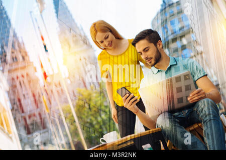 Der bärtige Mann sitzt mit zwei Gadgets, während seine Freundin in der Nähe von Stockfoto