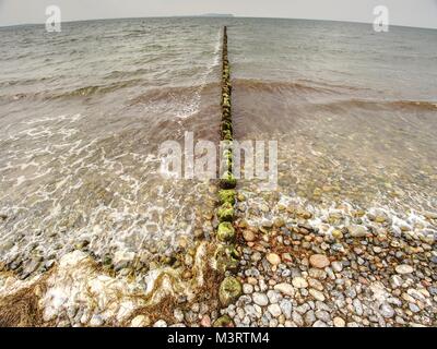 Alte hölzerne Wellenbrecher auf einem Ufer der Ostsee. Weiß schäumende Wasser Steine am Strand. Stockfoto