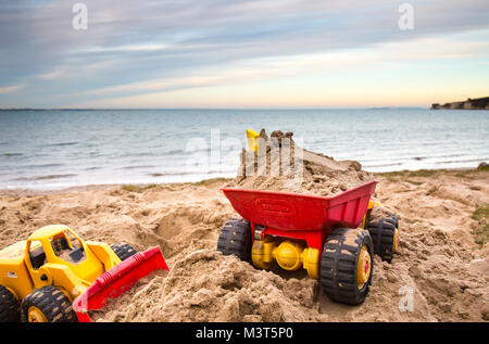 Nahaufnahme von Spielzeug Bagger und Lkw's ein Kind im Sand aufgegeben, auf einer verlassenen UK Strand. Am frühen Abend Sonnenlicht erzeugt Farben über Himmel. Stockfoto