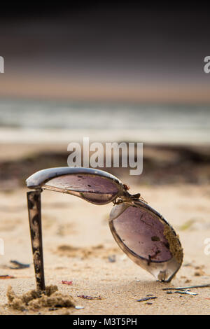 In der Nähe von Broken Sonnenbrille am Strand verlassen, im Sand unter Treibholz Schutt stecken gewaschen an Land durch die Gezeiten. Weichzeichner Strand & Himmel Hintergrund. Stockfoto