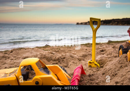 Nahaufnahme von Spielzeug Bagger & eines Kindes Spaten in den Sand aufgegeben, auf einer verlassenen UK Strand. Am frühen Abend Sonnenlicht erzeugt Farben über Himmel. Stockfoto